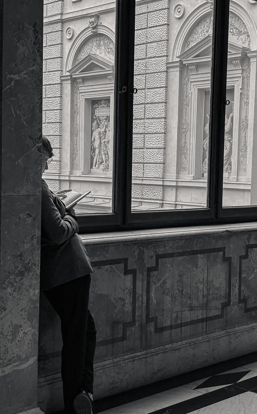 Figure reading by a museum window, ornate facade opposite